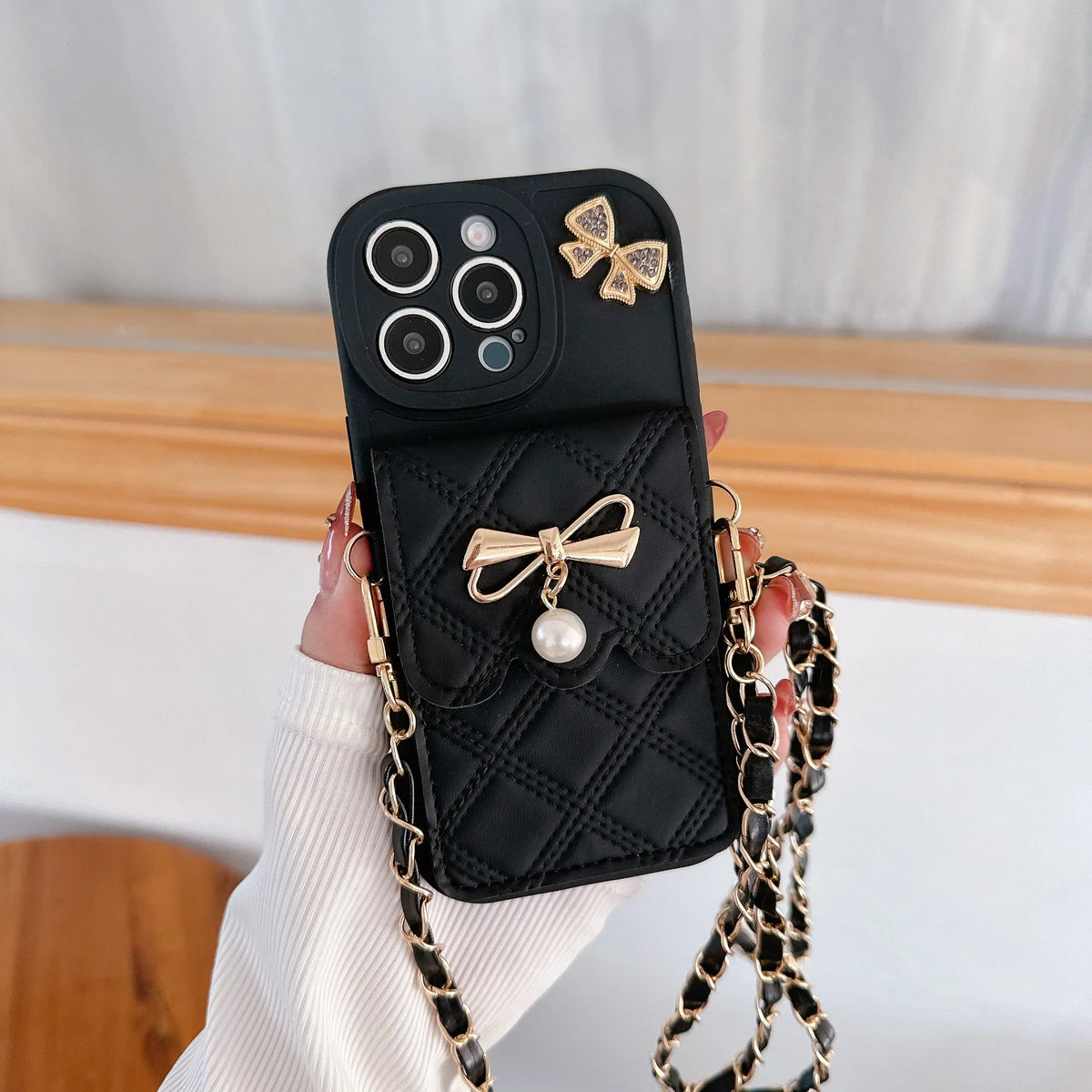 Black phone case with gold bows and a chain strap held by a hand.