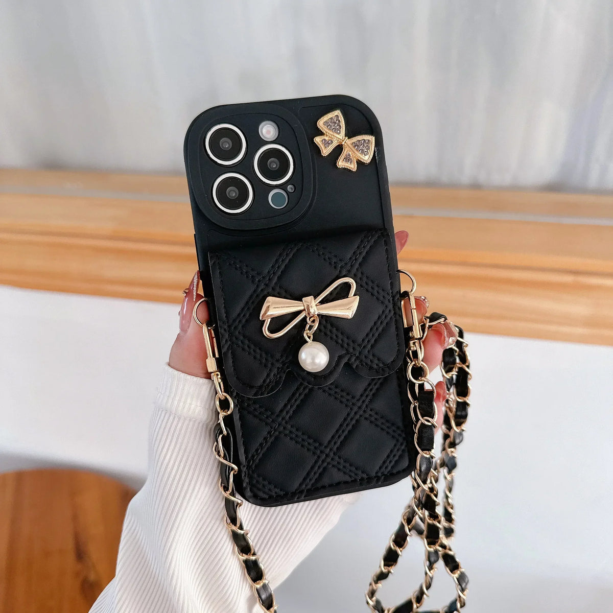 Black phone case with gold bows and a chain strap held by a hand.