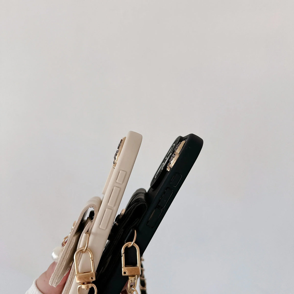 Two slim smartphone cases held in a hand against a plain light background. One case is cream/off-white and the other is black, both with gold-tone hardware and a matching gold ring detail.