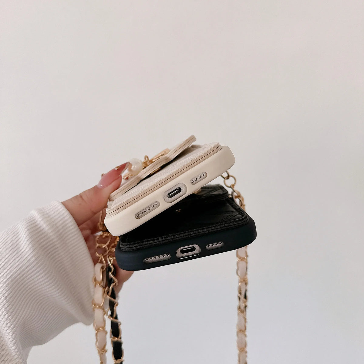 A hand wearing a white ribbed long-sleeve top holds two slim phone cases stacked vertically. The top case is cream with a gold chain strap and decorative gold hardware, and the bottom case is black with a matching black-and-gold chain strap.